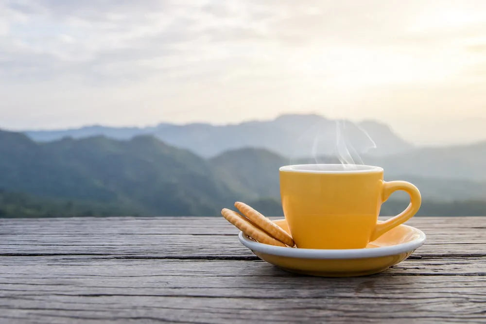 A white cup of hot espresso coffee mugs placed with cookies on a wooden floor with morning fog and mountains with sunlight background